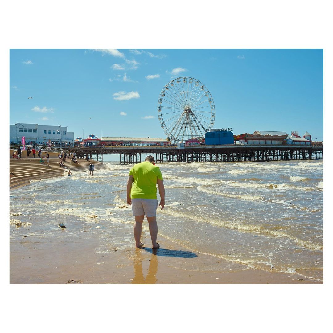 A man wearing a neon green t-shirt and grey shorts stands on a sandy beach near the water facing a large pier and ferris wheel.