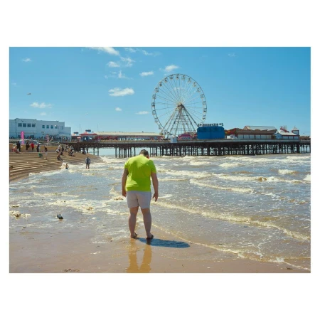 A man wearing a neon green t-shirt and grey shorts stands on a sandy beach near the water facing a large pier and ferris wheel.