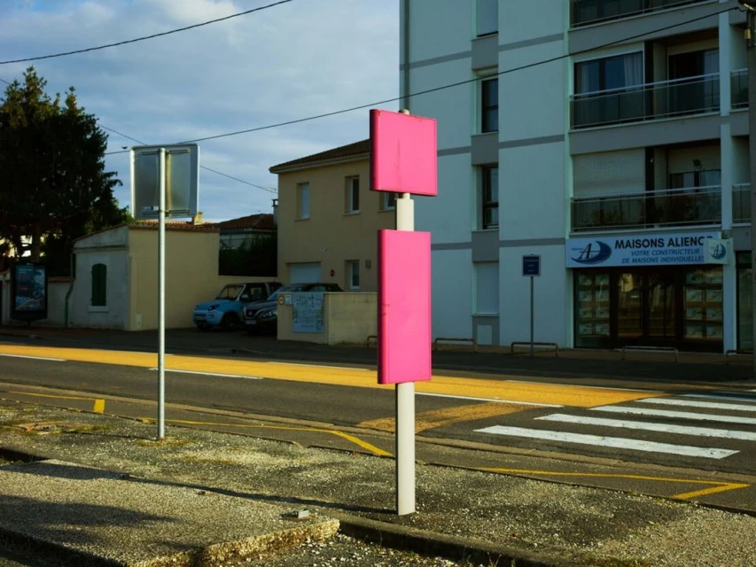 Pink signs stand beside a road with crosswalk and yellow line, near buildings and trees, captured in a daytime outdoor setting.