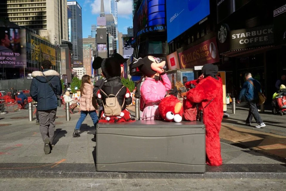 Costumed performers, including Mickey Mouse and Elmo, stand on a sidewalk in Times Square, New York City, surrounded by pedestrians and billboards.