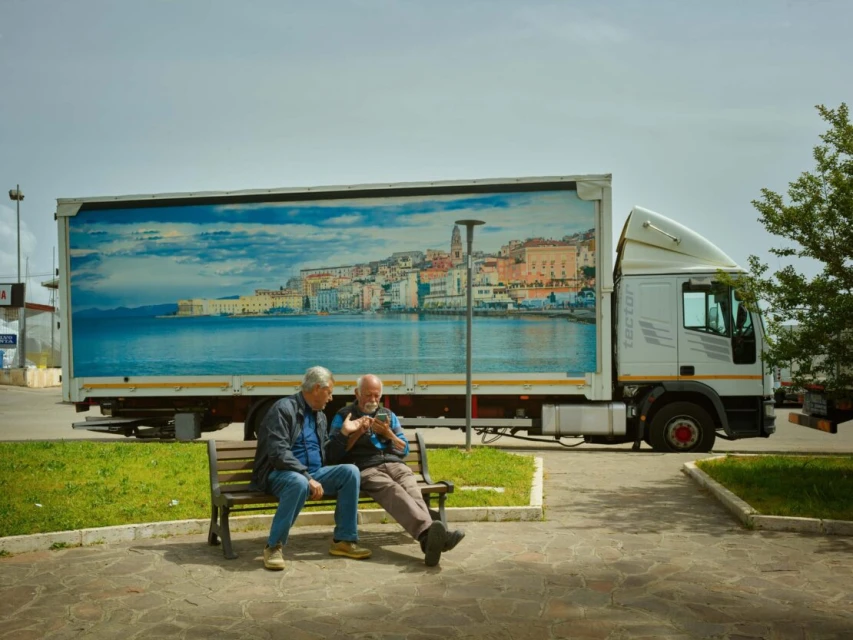 Two elderly men sit on a park bench, looking at a phone in front of a large truck with an image of a coastal town and blue sky.
