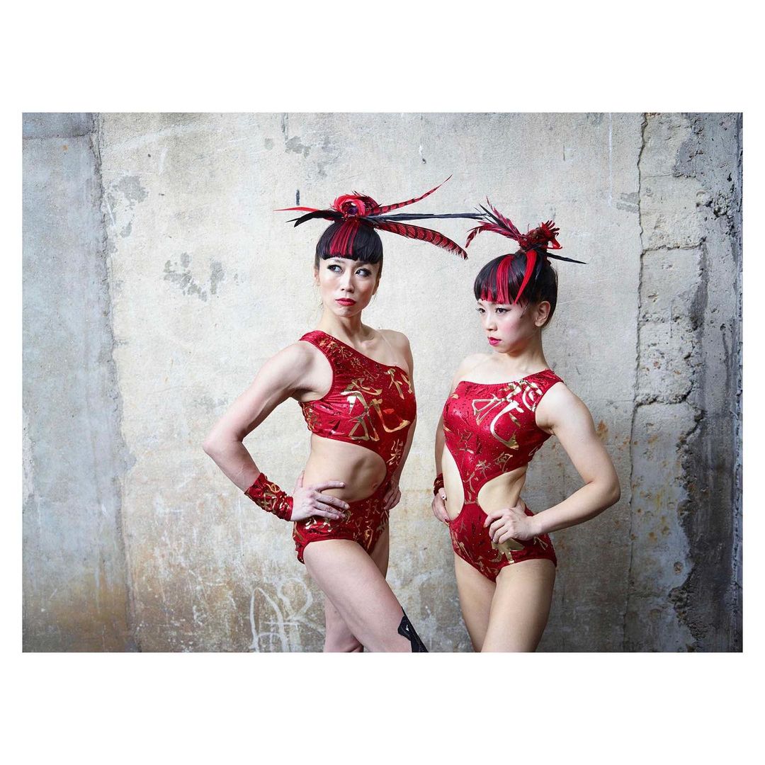 Two Asian women in red leotards and feathered headpieces pose in front of a textured grey wall, offering a striking and theatrical visual.
