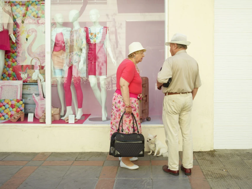 Elderly couple stands on a sidewalk with a small dog near a shop window displaying mannequins and colorful merchandise in the daytime.