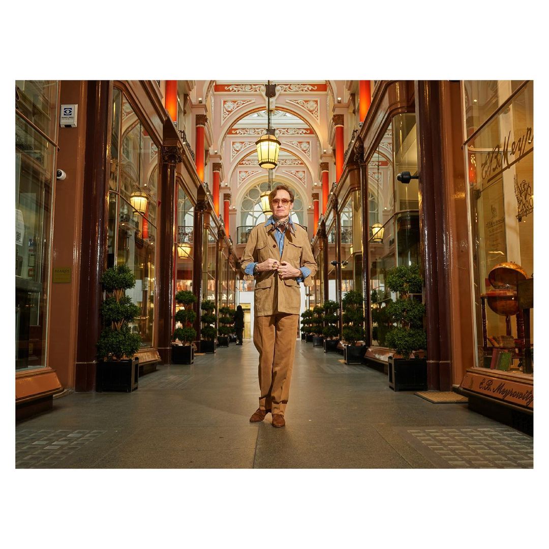 Adult male in a light brown suit and glasses poses indoors in an arcade with decorated arches, lanterns, and shopfronts.