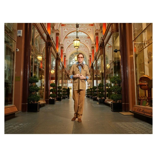 Adult male in a light brown suit and glasses poses indoors in an arcade with decorated arches, lanterns, and shopfronts.