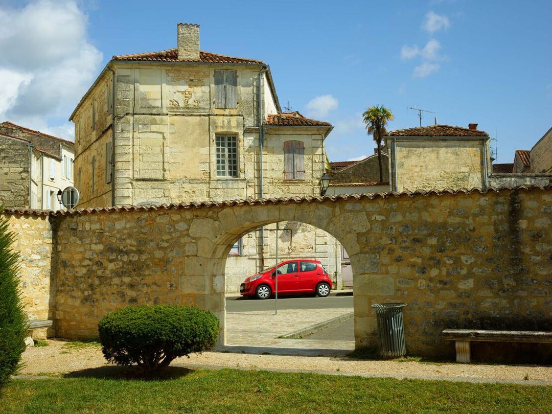 Exterior shot of a quaint stone building with an arched entryway, showcasing a glimpse of a red car on the street. Blue sky and greenery.
