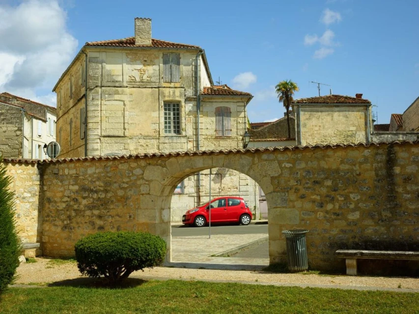 Exterior shot of a quaint stone building with an arched entryway, showcasing a glimpse of a red car on the street. Blue sky and greenery.