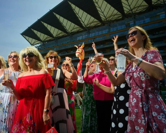 Group of women celebrating at an outdoor event on a sunny day, with drinks and a toy horse, and a large building in the background.