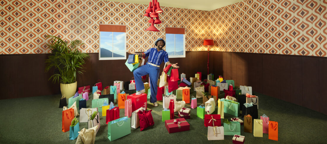 Cheerful African-American man stands amidst shopping bags in a retro-styled room, dressed in blue, with a patterned wallpaper and green carpet.