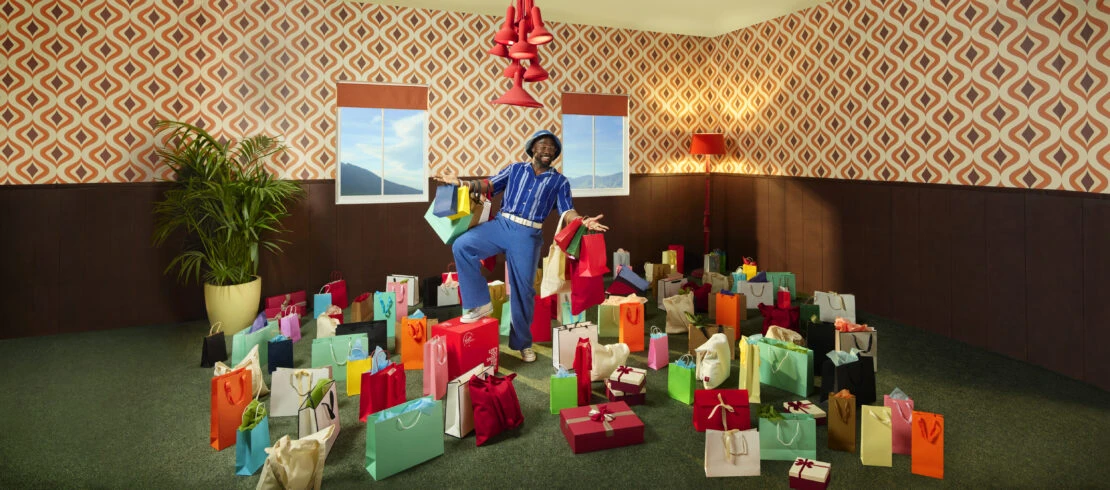 Cheerful African-American man stands amidst shopping bags in a retro-styled room, dressed in blue, with a patterned wallpaper and green carpet.