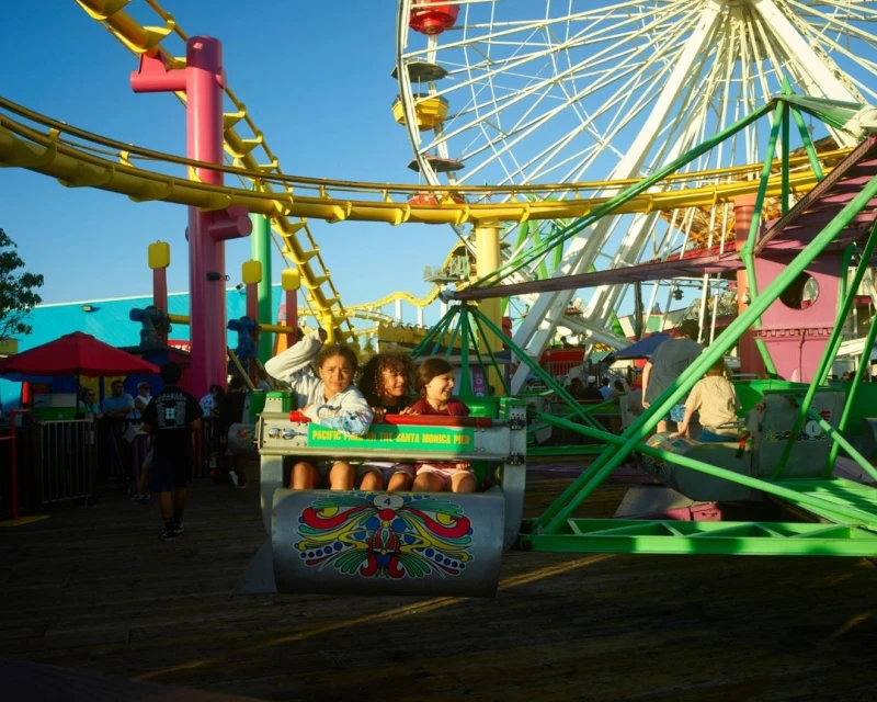 Three children ride an amusement park attraction with a roller coaster, Ferris wheel, and boardwalk visible in the background under a blue sky.