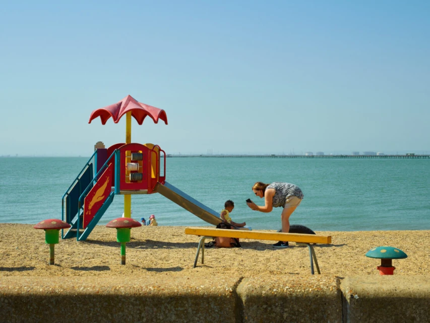 Woman with cell phone photographs a child near colorful playground on sandy beach with ocean background under a clear sky, daytime.
