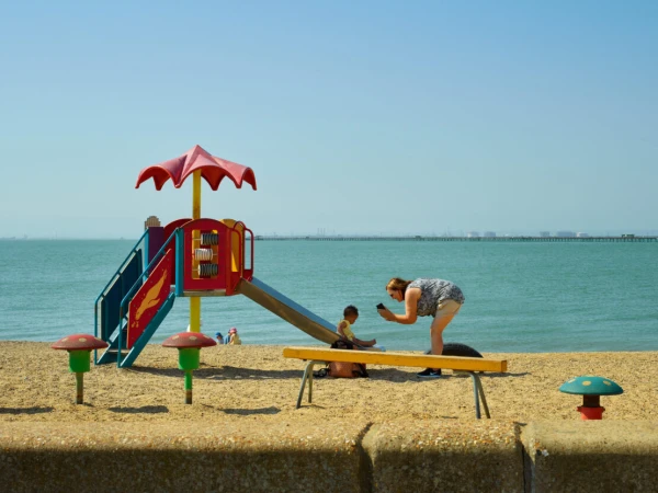 Woman with cell phone photographs a child near colorful playground on sandy beach with ocean background under a clear sky, daytime.