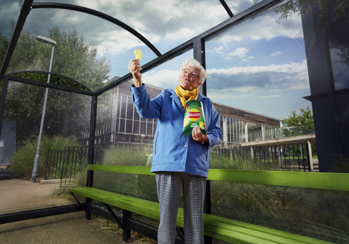 Elderly woman holding a crisp up to inspect it inside a glass bus shelter holding a Walkers crisps packet