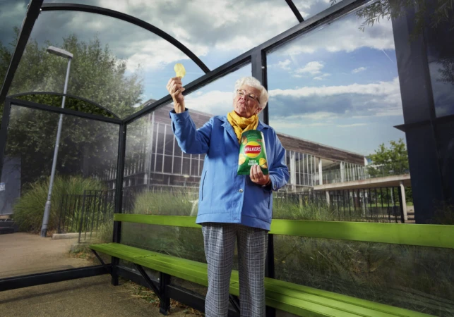 Elderly woman holding a crisp up to inspect it inside a glass bus shelter holding a Walkers crisps packet