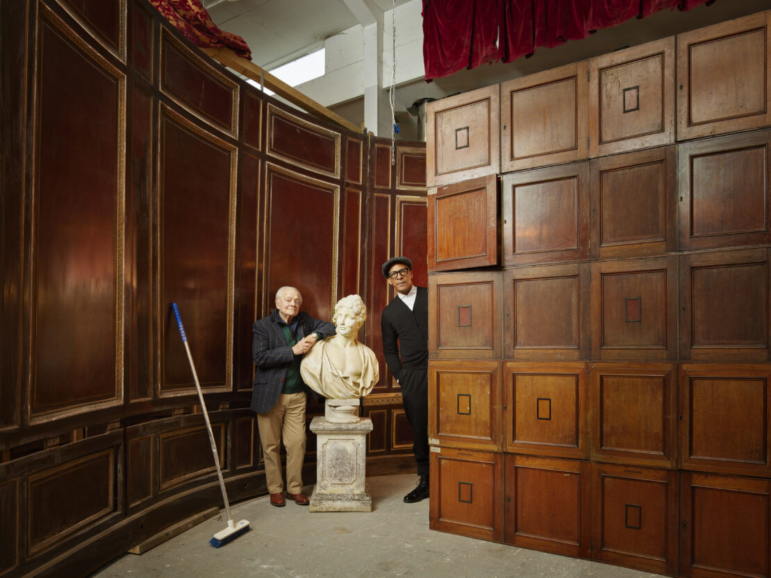 Two men pose beside a bust amid paneled walls and red curtains, with a broom propped against the wall, creating a staged appearance.