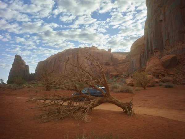 Scenic view of a desert landscape featuring a blue car parked near a weathered tree, set against red rock formations and a cloudy sky.