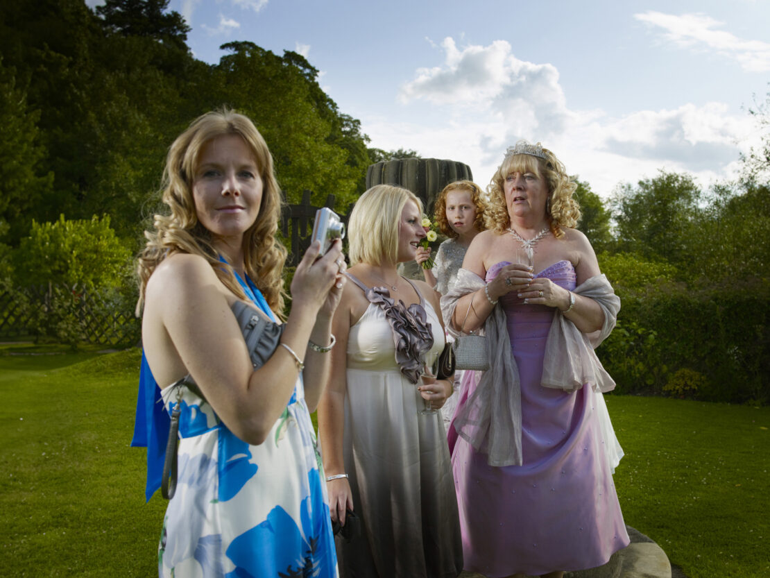 Group of women in formal dresses outdoors, one taking a photo with a compact camera, with lush green background and overcast sky.
