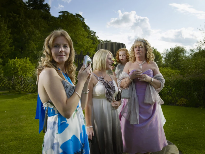 Group of women in formal dresses outdoors, one taking a photo with a compact camera, with lush green background and overcast sky.