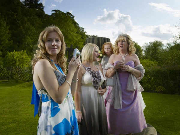 Group of women in formal dresses outdoors, one taking a photo with a compact camera, with lush green background and overcast sky.