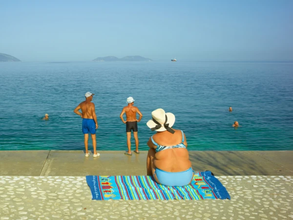Woman in blue bikini and wide-brim hat sitting on a striped towel by the ocean, with two standing men and swimmers nearby.