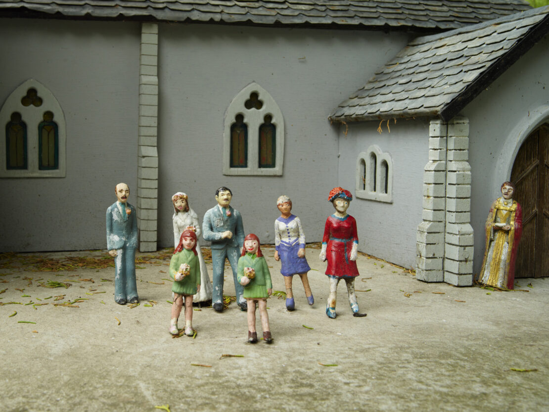 Miniature wedding scene with figurines of a bride, groom, guests and officiant outside a grey-stone church under a cloudy sky.