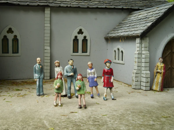 Miniature wedding scene with figurines of a bride, groom, guests and officiant outside a grey-stone church under a cloudy sky.