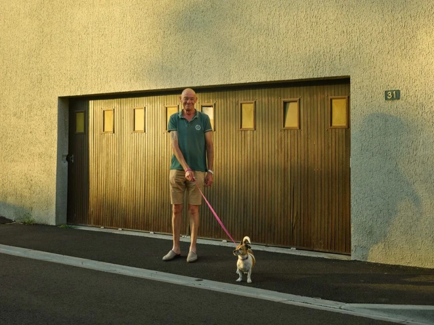 Elderly man walks his small dog on a leash in front of a modern building with a brown garage door on a sunny day.