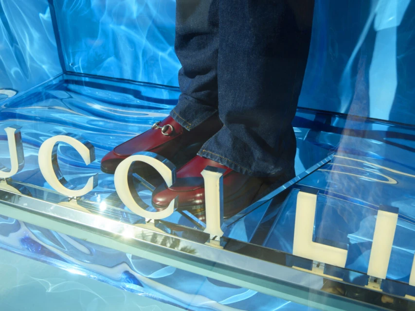 Gucci loafers on a reflective blue platform with the 'GUCCI' logo visible in a studio environment, promoting fashion and luxury.