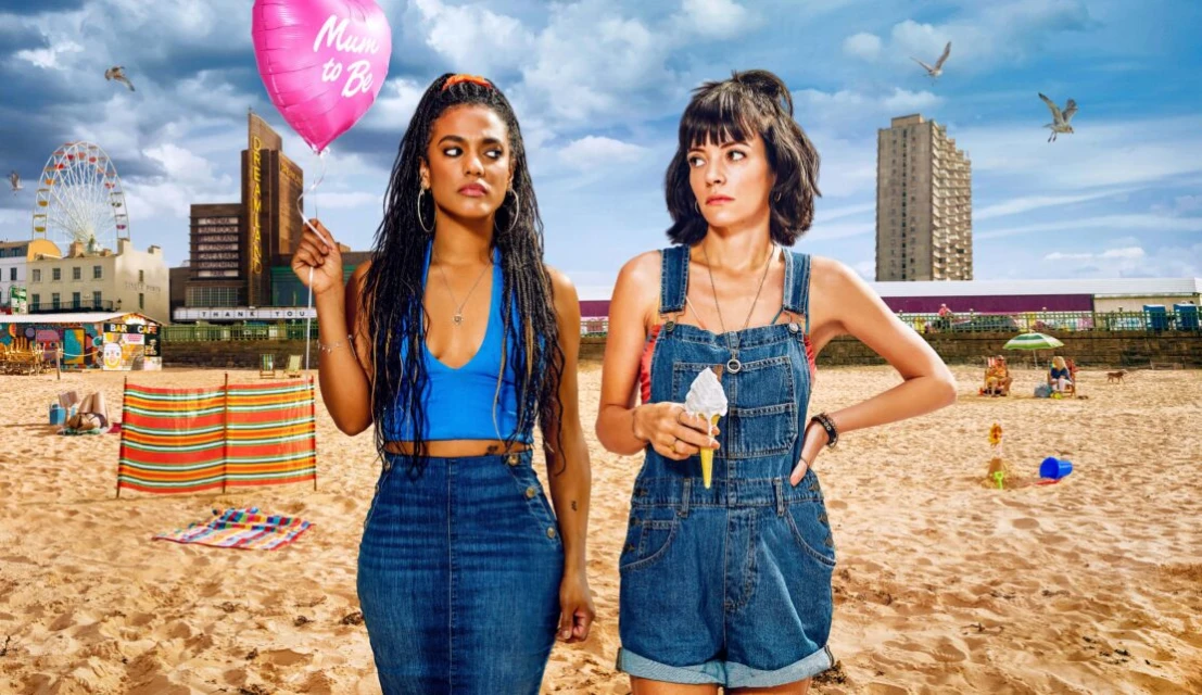 Two women stand on a sunny beach with a Ferris wheel in the background. One holds an ice cream cone and the other holds a balloon.