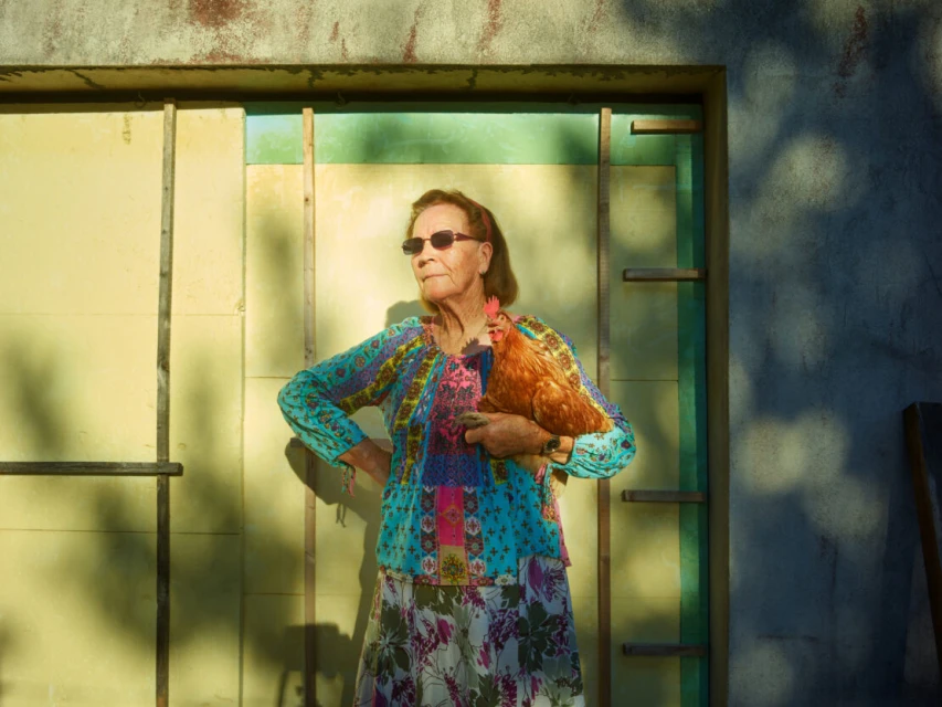 Elderly woman with sunglasses holds a brown hen in front of a weathered building, creating a warm and nostalgic outdoor portrait.