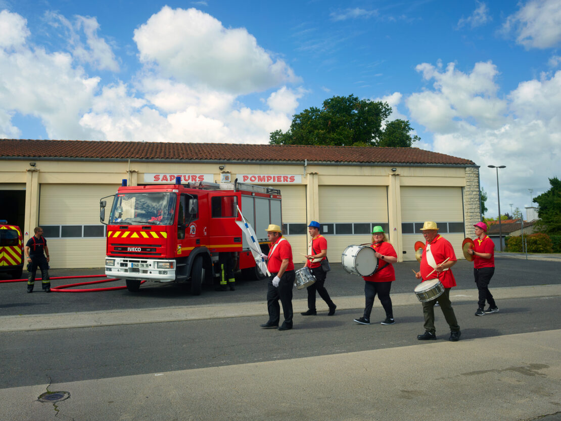 A group of musicians dressed in red marching in a parade in front of a fire station, with a fire engine, under a blue sky with clouds.