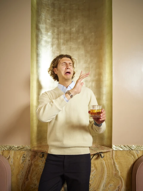 Young man with curly hair pulls a face with hand raised, holding a glass of amber alcohol against a gold backdrop indoors.