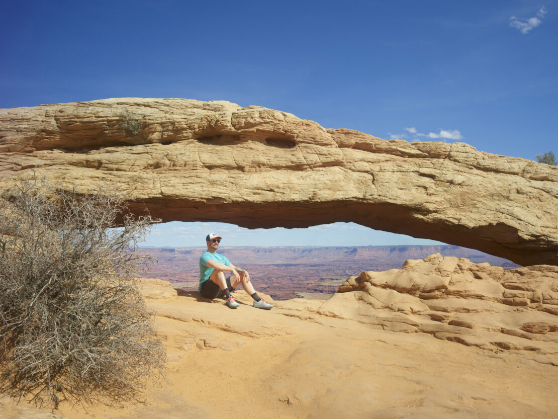 Man sits beneath Mesa Arch in Canyonlands National Park, Utah. Clear blue sky above desert rock formation frames scenic view.