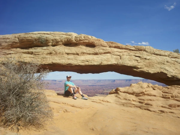 Man sits beneath Mesa Arch in Canyonlands National Park, Utah. Clear blue sky above desert rock formation frames scenic view.