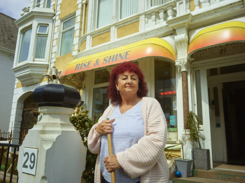 Woman stands in front of the Rise & Shine bed and breakfast, holding a broom. The building is cream-colored with yellow and orange awnings.