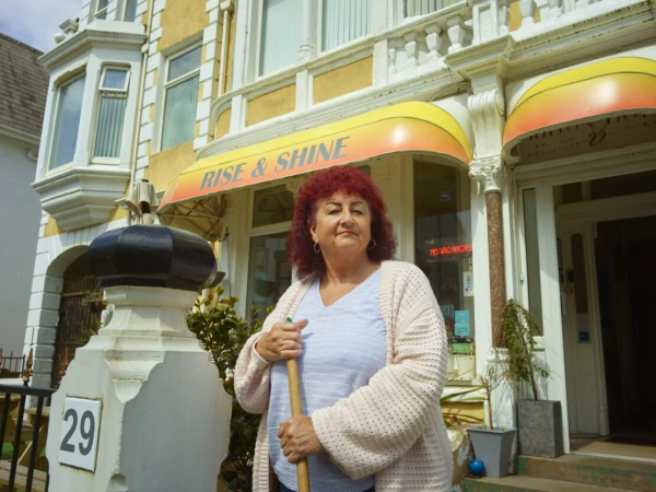 Woman stands in front of the Rise & Shine bed and breakfast, holding a broom. The building is cream-colored with yellow and orange awnings.