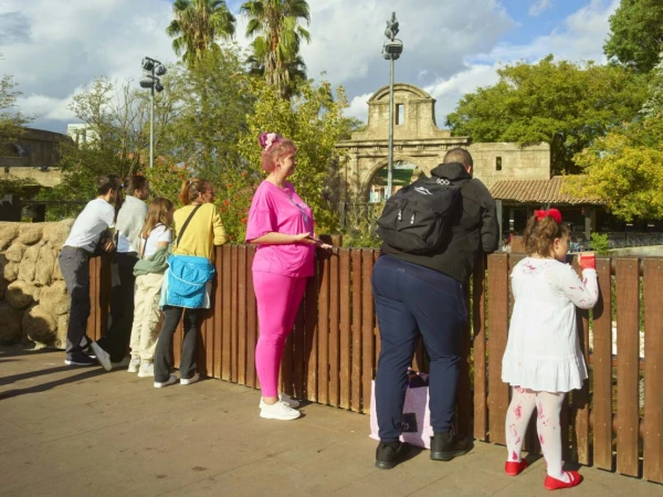 Group of people of varying ages stand along a wooden fence in an outdoor park setting, overlooking an enclosure, on a sunny day.
