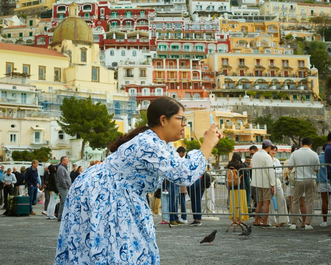 Woman in blue patterned dress takes photo of Positano, Italy, with apartment buildings, people, and birds in outdoor daylight.