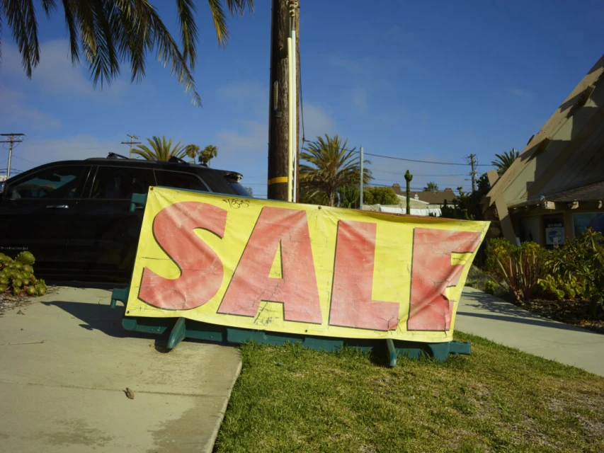 Outdoor shot of a yellow sale banner with large red letters on a green grass lawn next to a sidewalk and a black SUV.