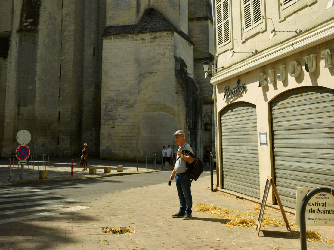 An adult male stands on a city street near a shop with metal shutters and a large stone building in the background on a sunny day.