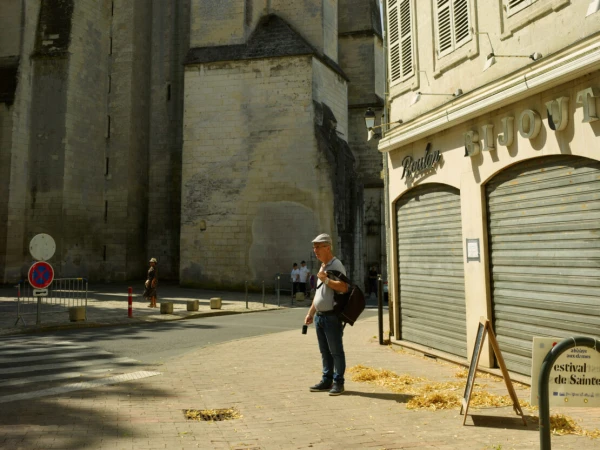 An adult male stands on a city street near a shop with metal shutters and a large stone building in the background on a sunny day.