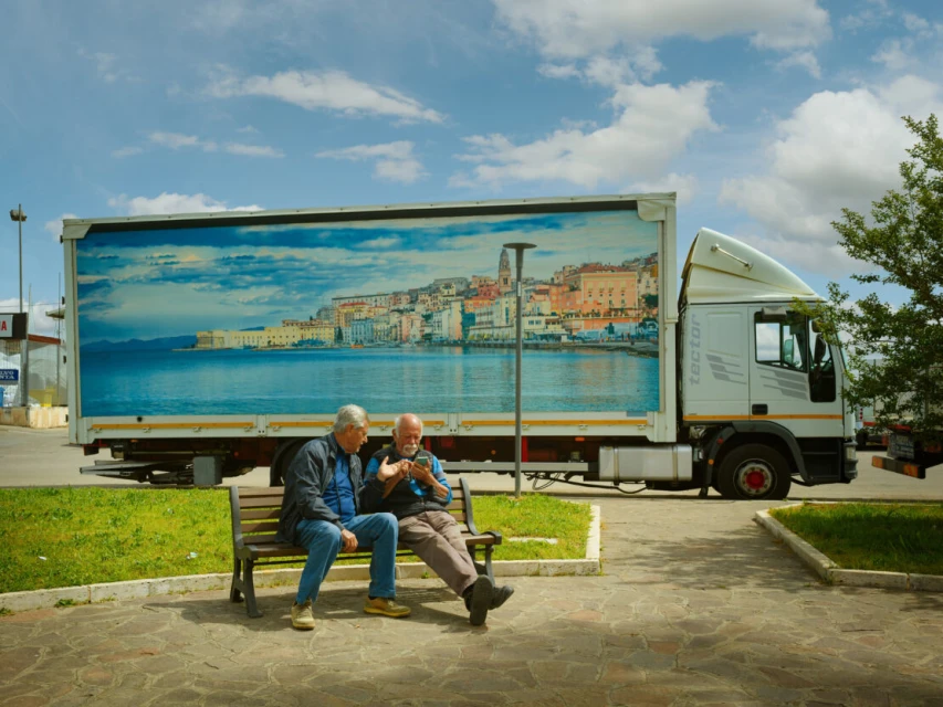 Two elderly men sit on a bench in an urban setting, with a large truck featuring a coastal city image parked behind them on a sunny day.