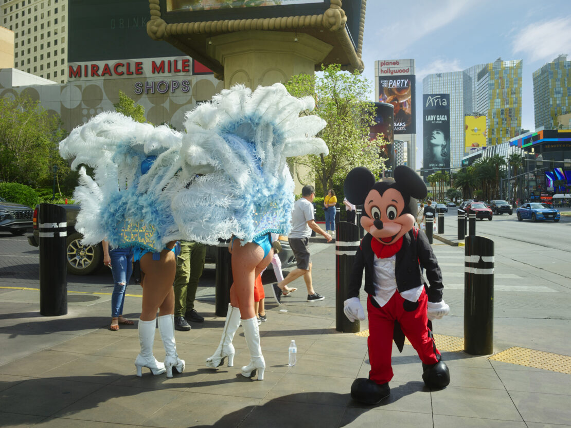 Mascot and showgirls perform on a Las Vegas sidewalk in front of hotels, shops, and billboards, creating a vibrant and lively street scene.