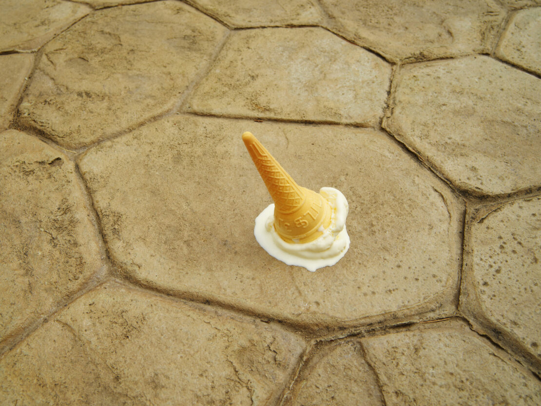 Fallen ice cream cone on a stone surface, vanilla ice cream melting around the cone, close-up shot of dessert mishap outdoors.