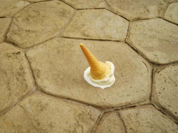 Fallen ice cream cone on a stone surface, vanilla ice cream melting around the cone, close-up shot of dessert mishap outdoors.