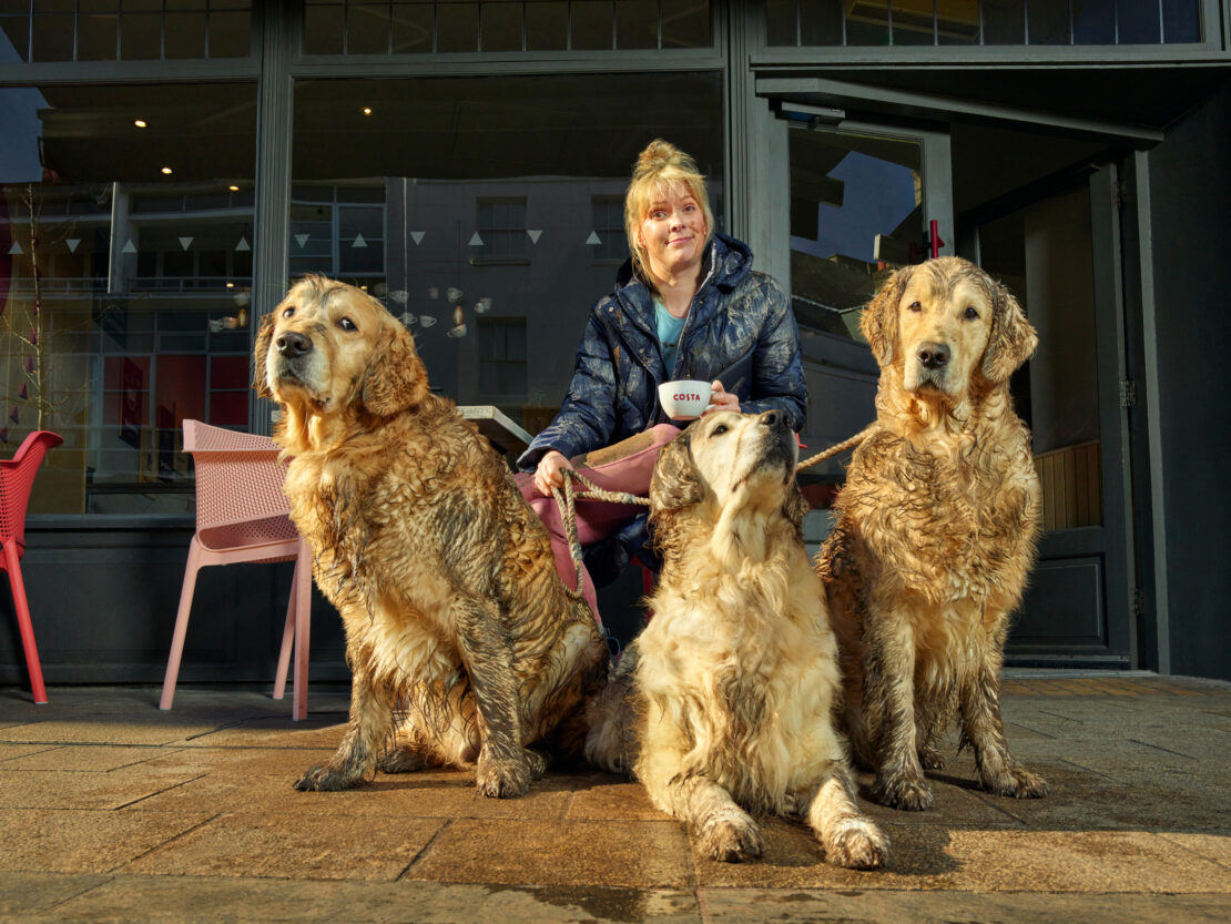Woman sits with three golden retrievers outside a Costa Coffee, one dog looks up to her while the others look off in opposite directions.