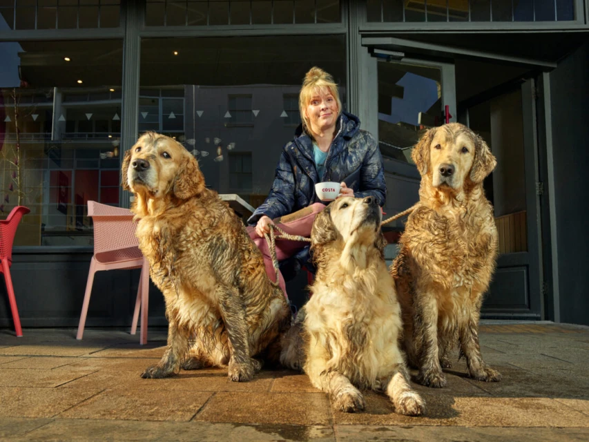 Woman sits with three golden retrievers outside a Costa Coffee, one dog looks up to her while the others look off in opposite directions.