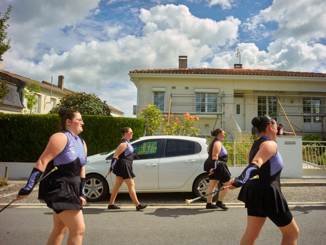 A group of baton twirlers walking in formation on a street lined with houses on a sunny day, wearing matching black and purple outfits.