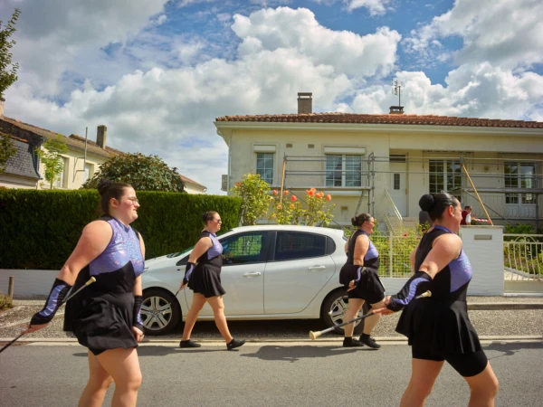 A group of baton twirlers walking in formation on a street lined with houses on a sunny day, wearing matching black and purple outfits.